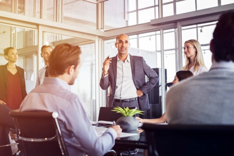 Professional setting around conference table. Several male and female coworkers sitting listening to male standing at head of table.